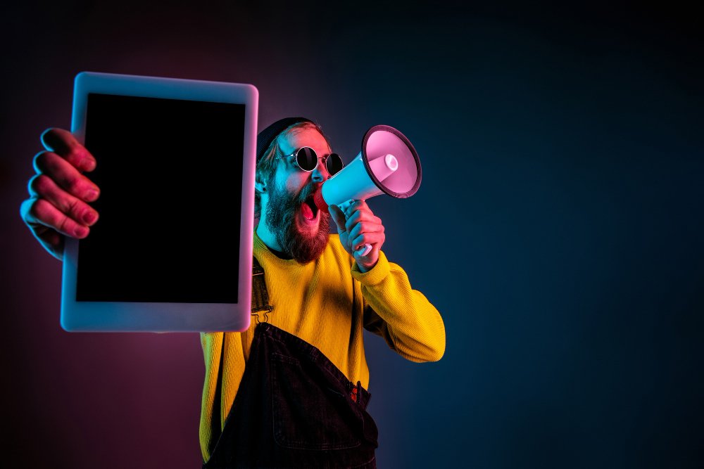 um homem barbudo de touca, vestindo uma blusa amarela e macacão preto e óculos escuros e na mão esquerda um megafone e na direita um tablet sem nenhum texto em sua tela fazendo alusão a um possível pregador cristão mas que prega sobre a internet, sobre os meios digitais.